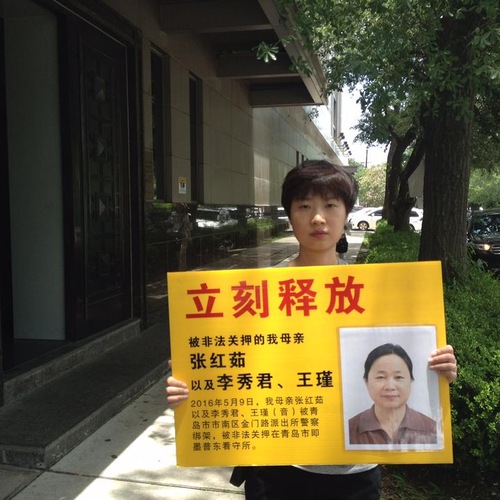 Anak perempuan Zhang memegang papan bertulisan, “Bebaskan segera ibu saya Zhang Hongru, yang ditangkap bersama dengan Li Xiujun dan Wang Jin pada 9 Mei 2016. Mereka sekarang ditahan di Pusat Penahanan Pudong.”